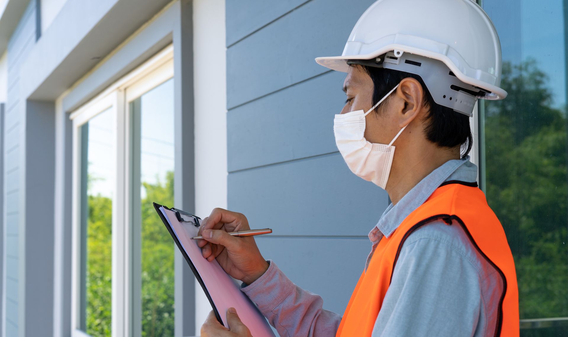 Worker in hard hat and mask taking notes outside a blue building