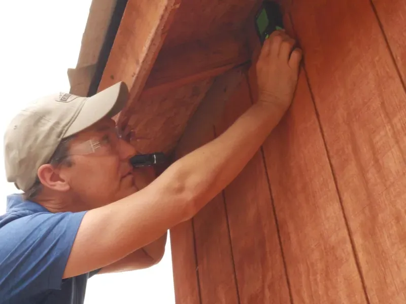 Man adjusting a wooden wall panel outdoors, wearing a cap and blue shirt