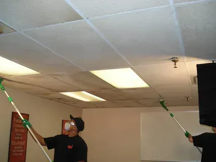People cleaning a ceiling with long poles and green tape in a room with fluorescent lights.