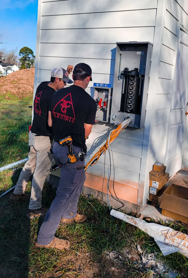 Two electricians working on an electrical panel outside a building. One holds a cable.