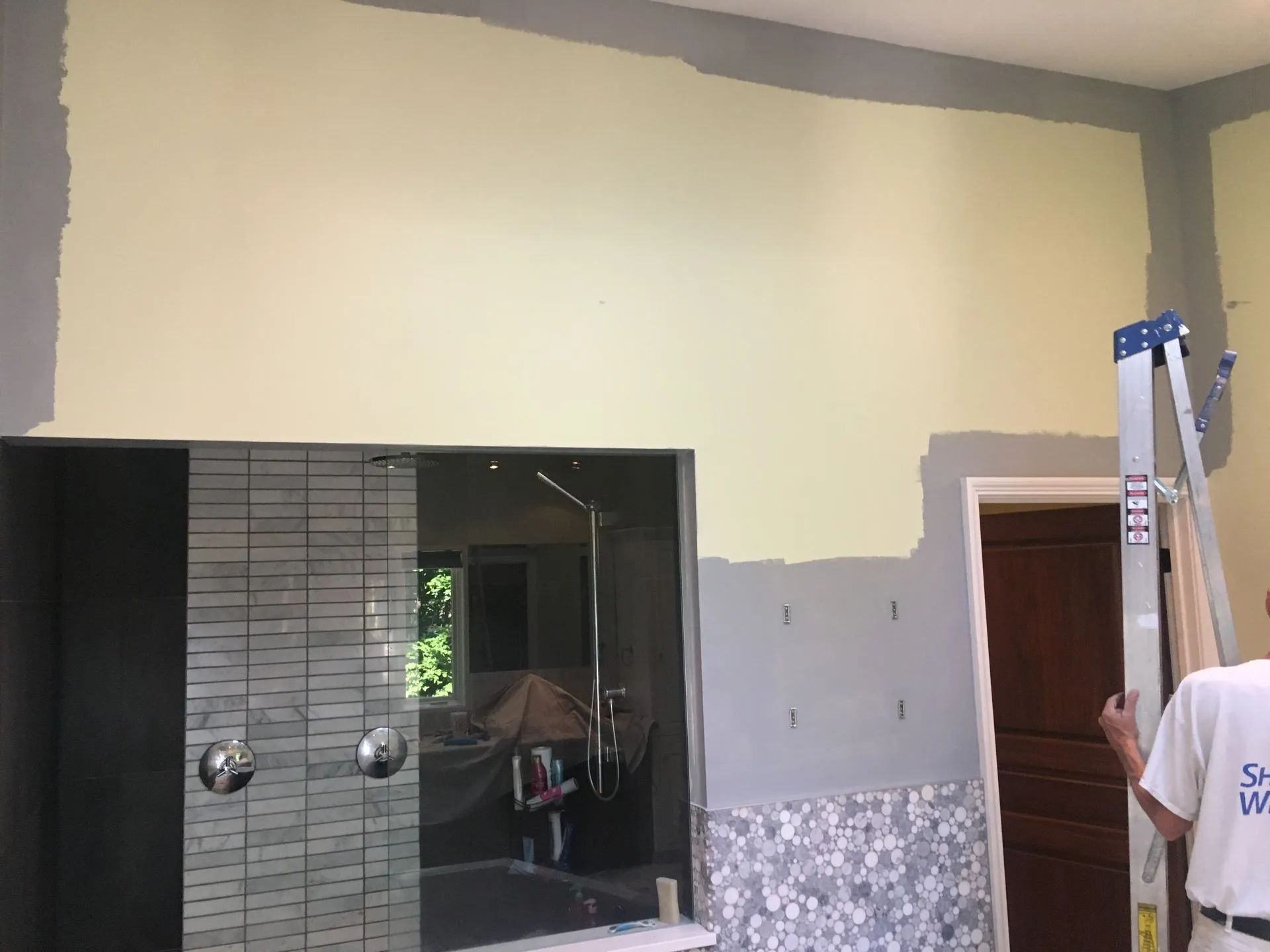 Indoor room with a tall cream wall being painted, with a ladder and worker at the right edge.