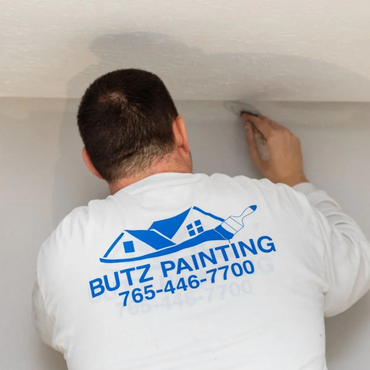 Painter in white shirt applying paint to a ceiling corner with a smoothing tool, showing a Butz Painting logo.