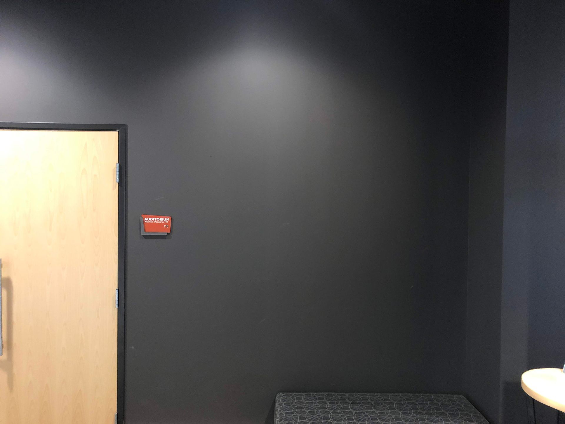 Empty dark room with black wall, wooden door, and a small bench in the corner.