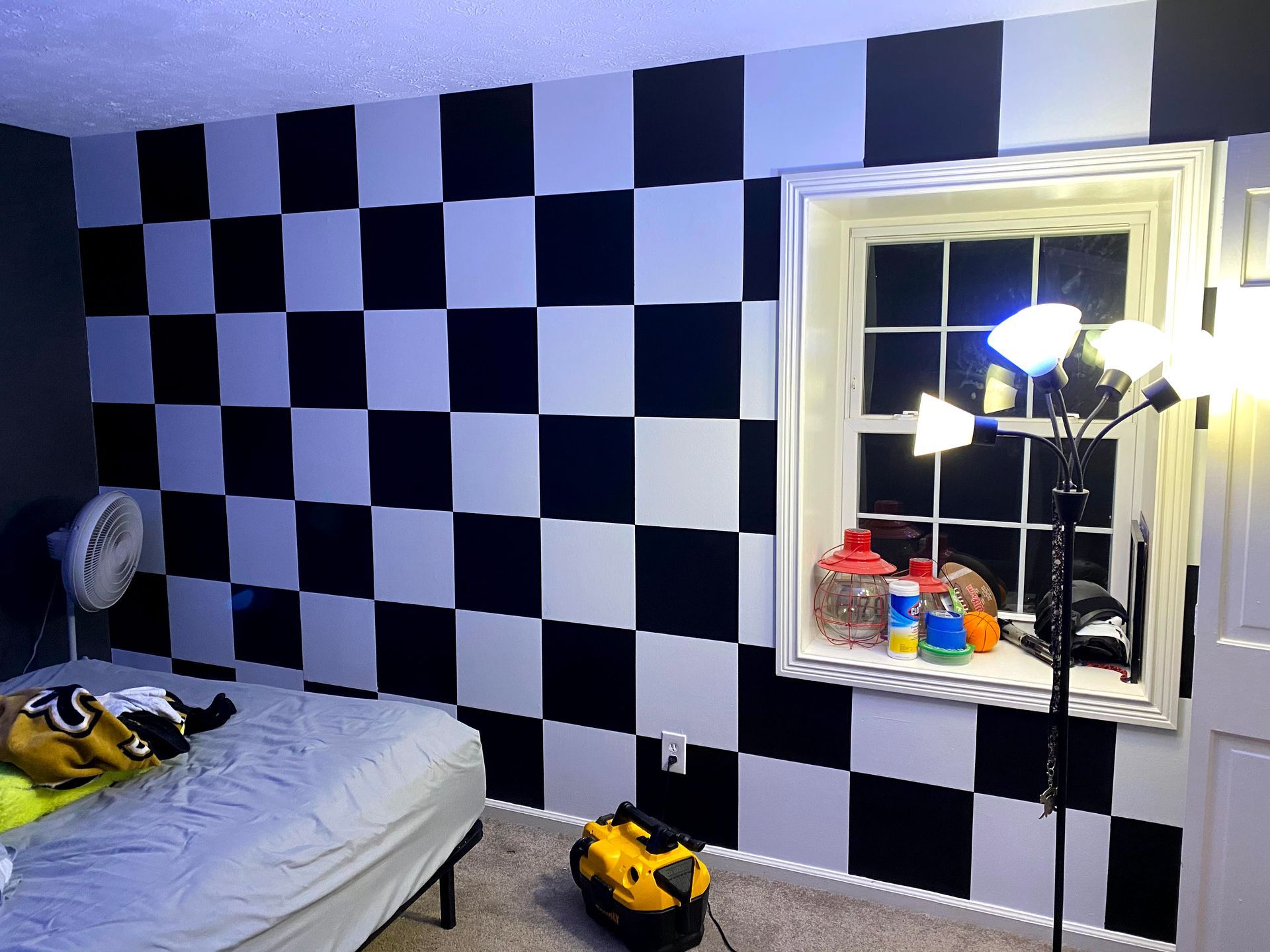 Bedroom with black-and-white checkerboard wall, bed, mirror, lamp, and toy cars on the floor
