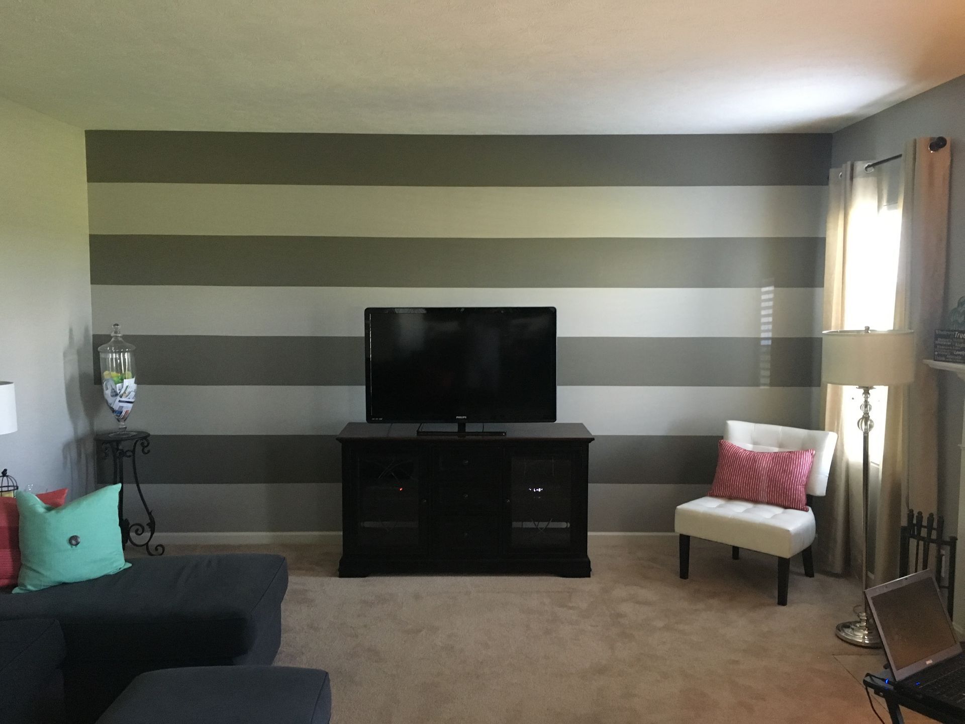 Living room with striped accent wall, TV on stand, couch, chair, and lamp with window light