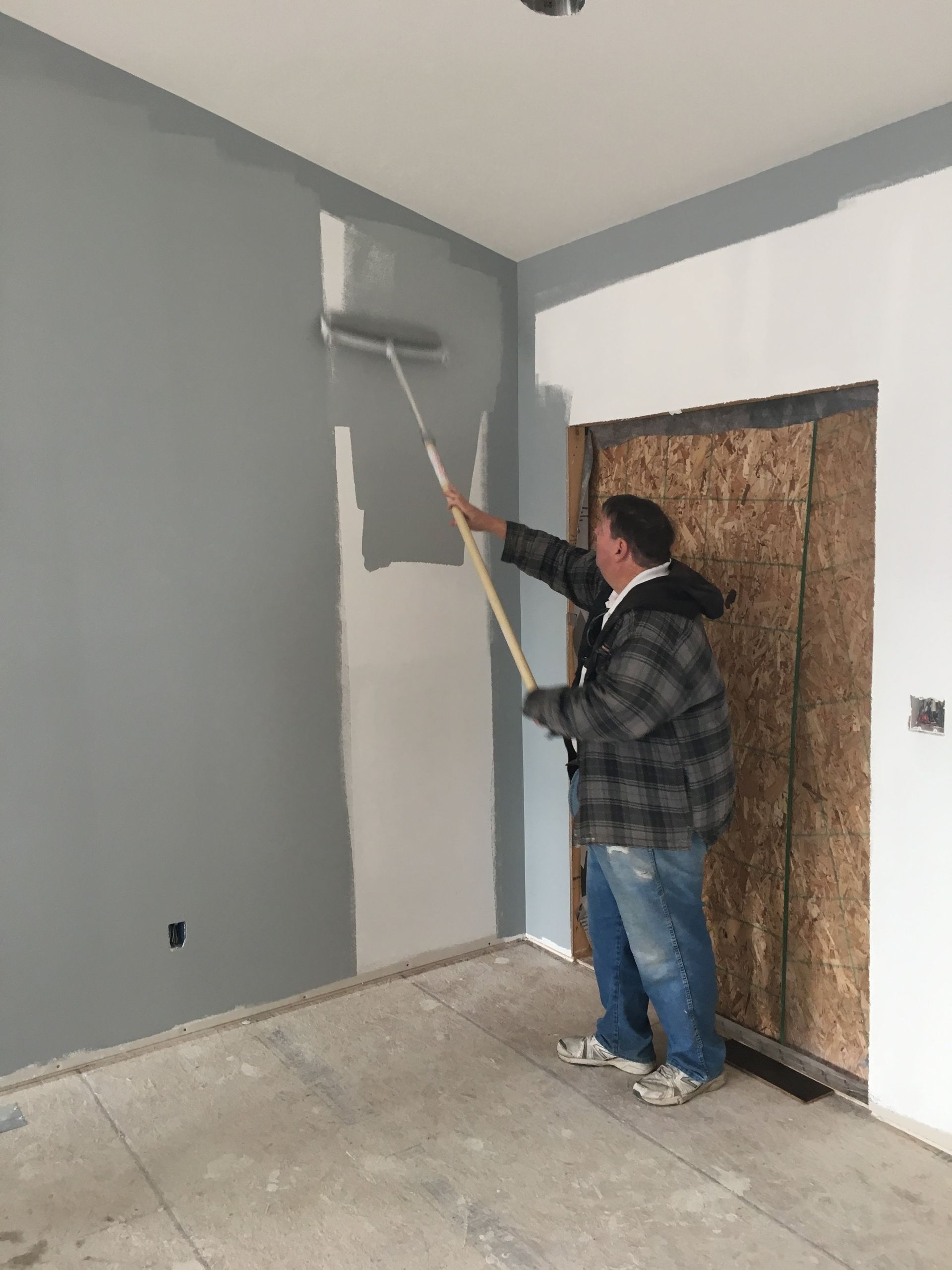 Person painting a gray wall with a roller in a nearly empty room with plywood-covered doorway.