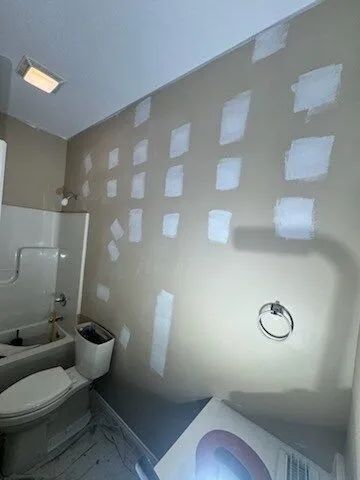 Small bathroom under renovation with patchy drywall, toilet, shower, and sink visible.