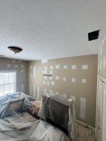 Living room with patchy beige walls, ceiling lights, and furniture covered in plastic for painting or ремонт