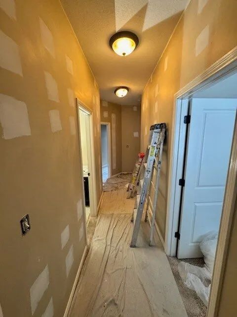 Narrow hallway under renovation with taped drywall, ladders, and a doorway on the right
