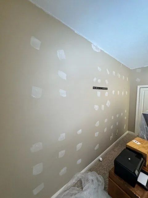 Drywalled beige room with white patch spots, a door, and a small black cabinet in the corner.