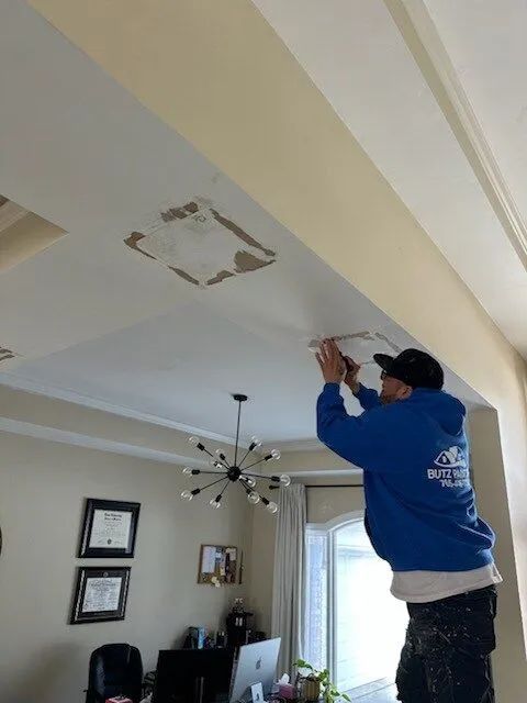 Person in blue shirt repairing a ceiling in a living room with a ladder and hanging light fixture