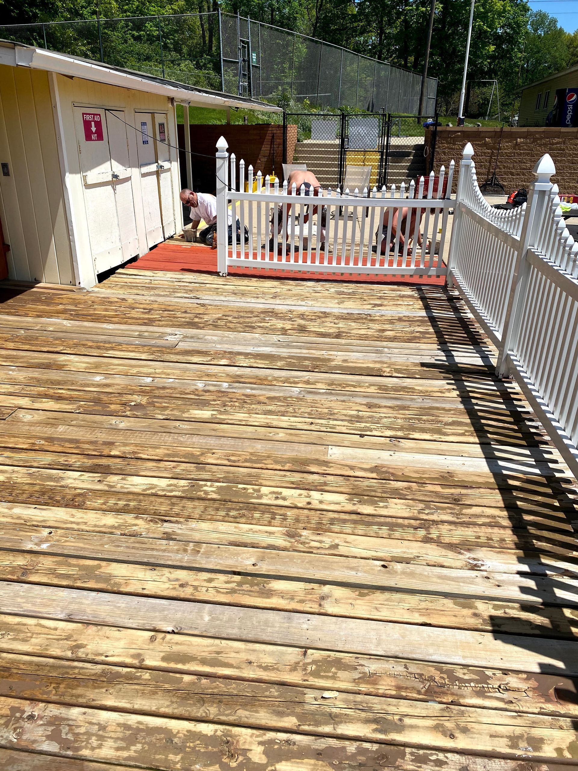 Sunlit wooden deck leading to a white picket fence and gate beside a small shed and trees