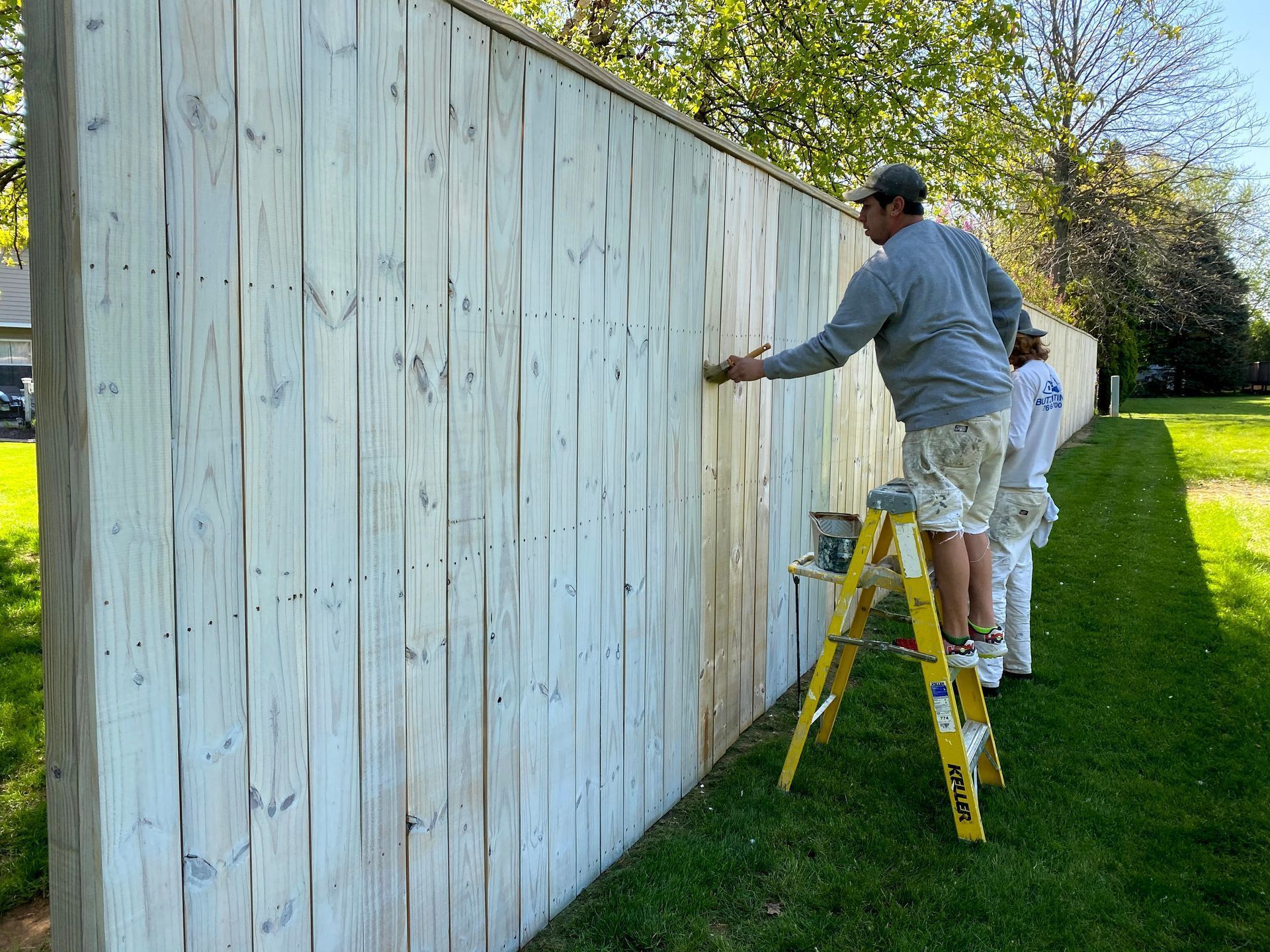 Two people painting a tall wooden fence outdoors on a sunny day