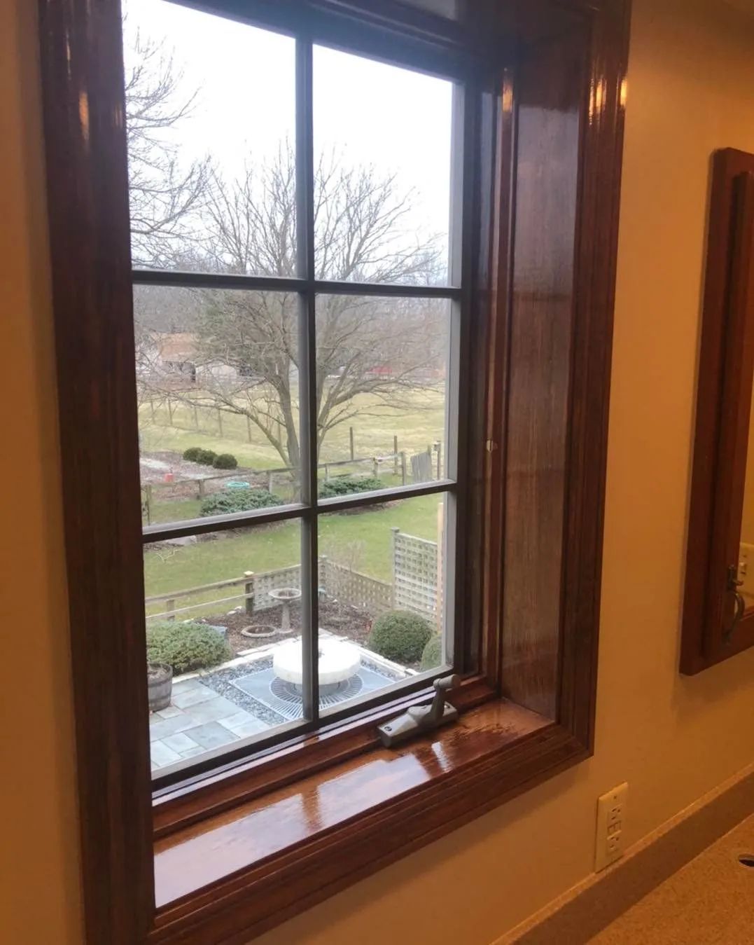 Wooden window overlooking a grassy yard with bare trees and a small patio below