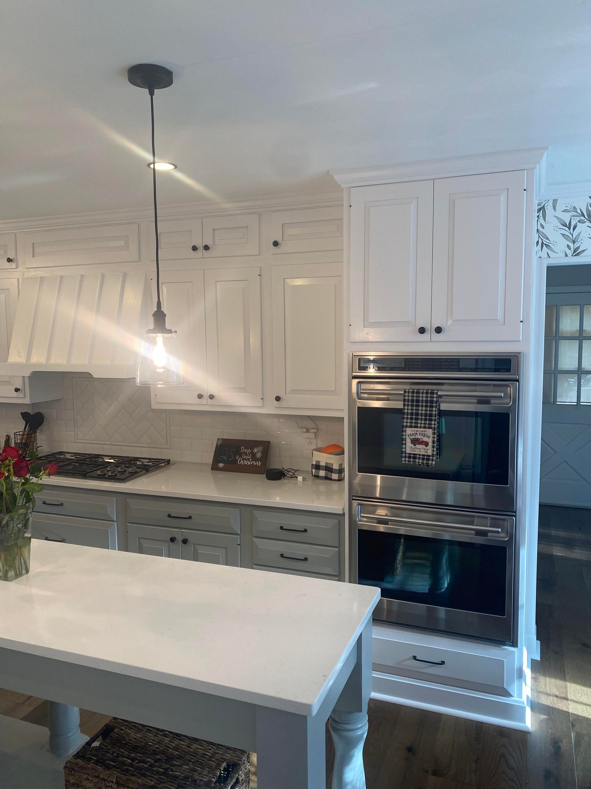 Bright white kitchen with island, pendant light, gray cabinets, double oven, and flowers on the counter