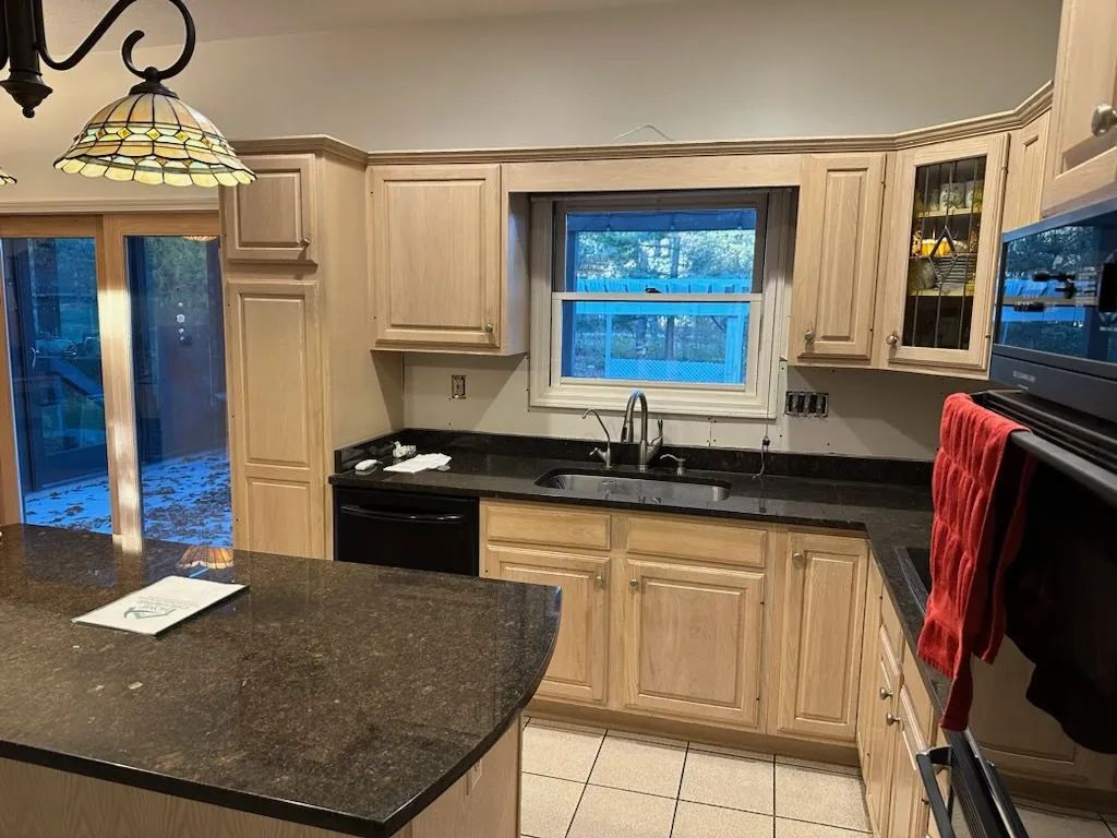 Kitchen with light wood cabinets, black countertops, and a window above the sink.