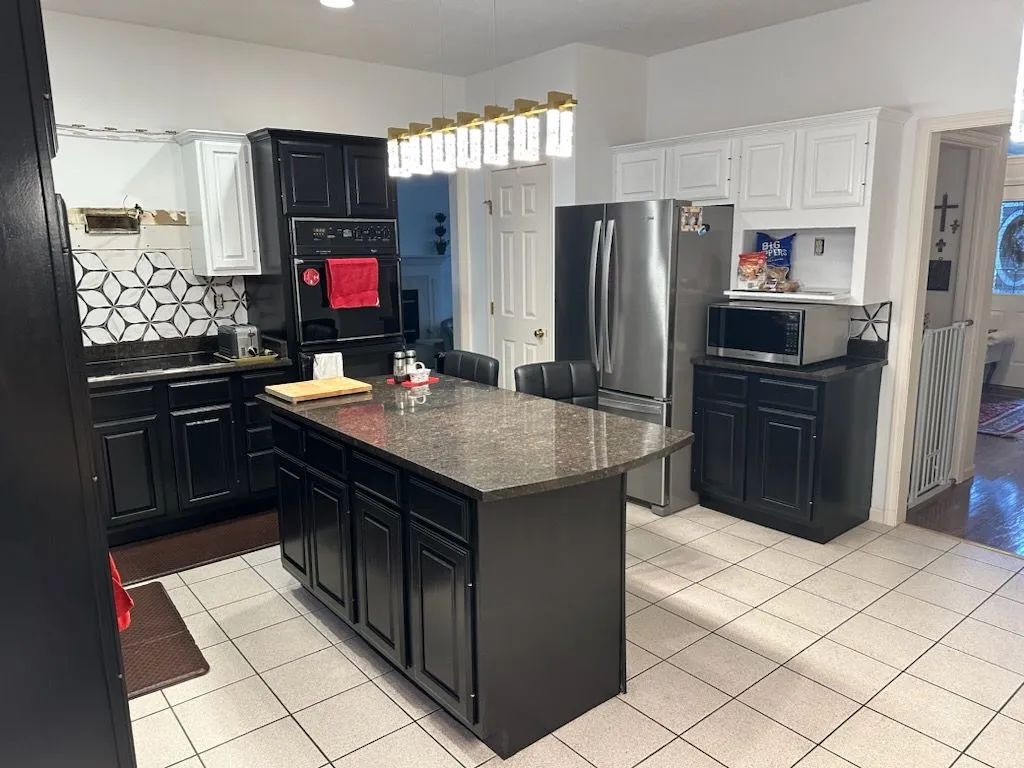 Modern kitchen with black cabinets, granite island, stainless steel fridge, and white tiled floor.
