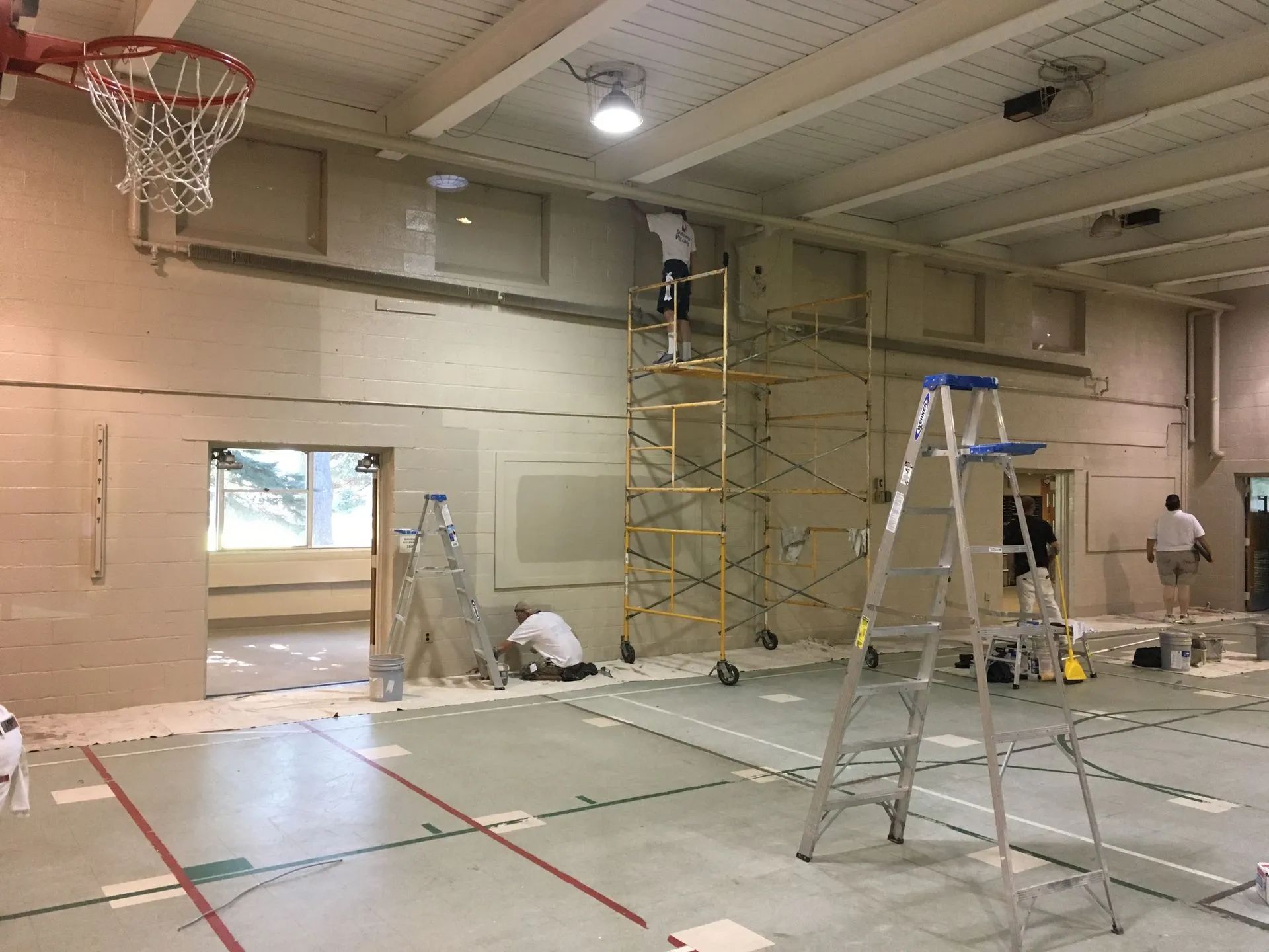 Workers painting a gym interior with ladders and scaffolding under a basketball hoop