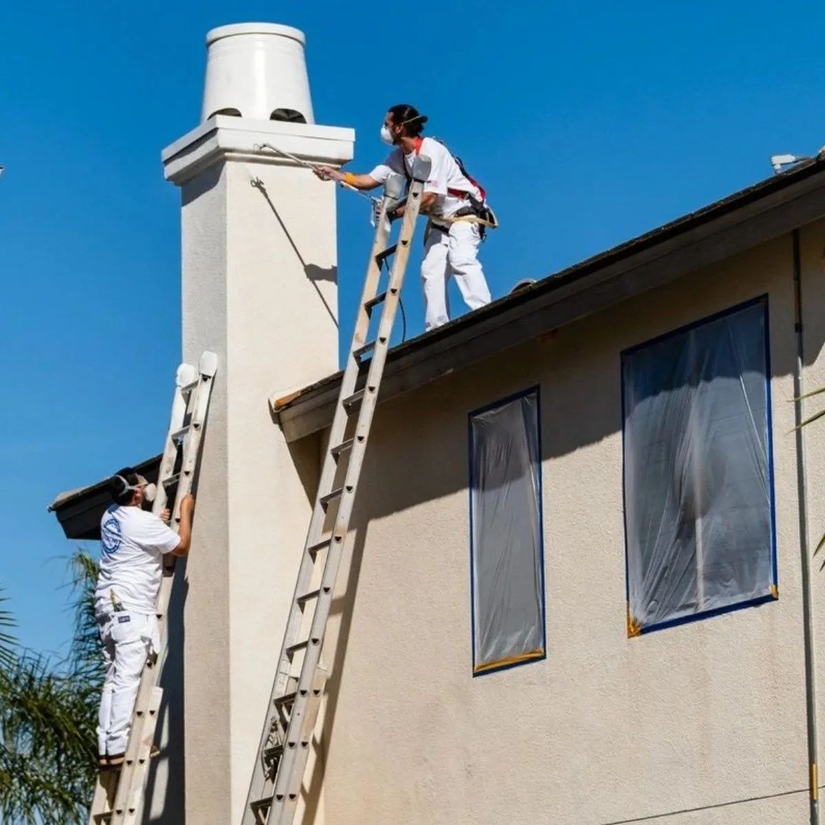 Two workers on ladders painting a house chimney and exterior wall under a bright blue sky