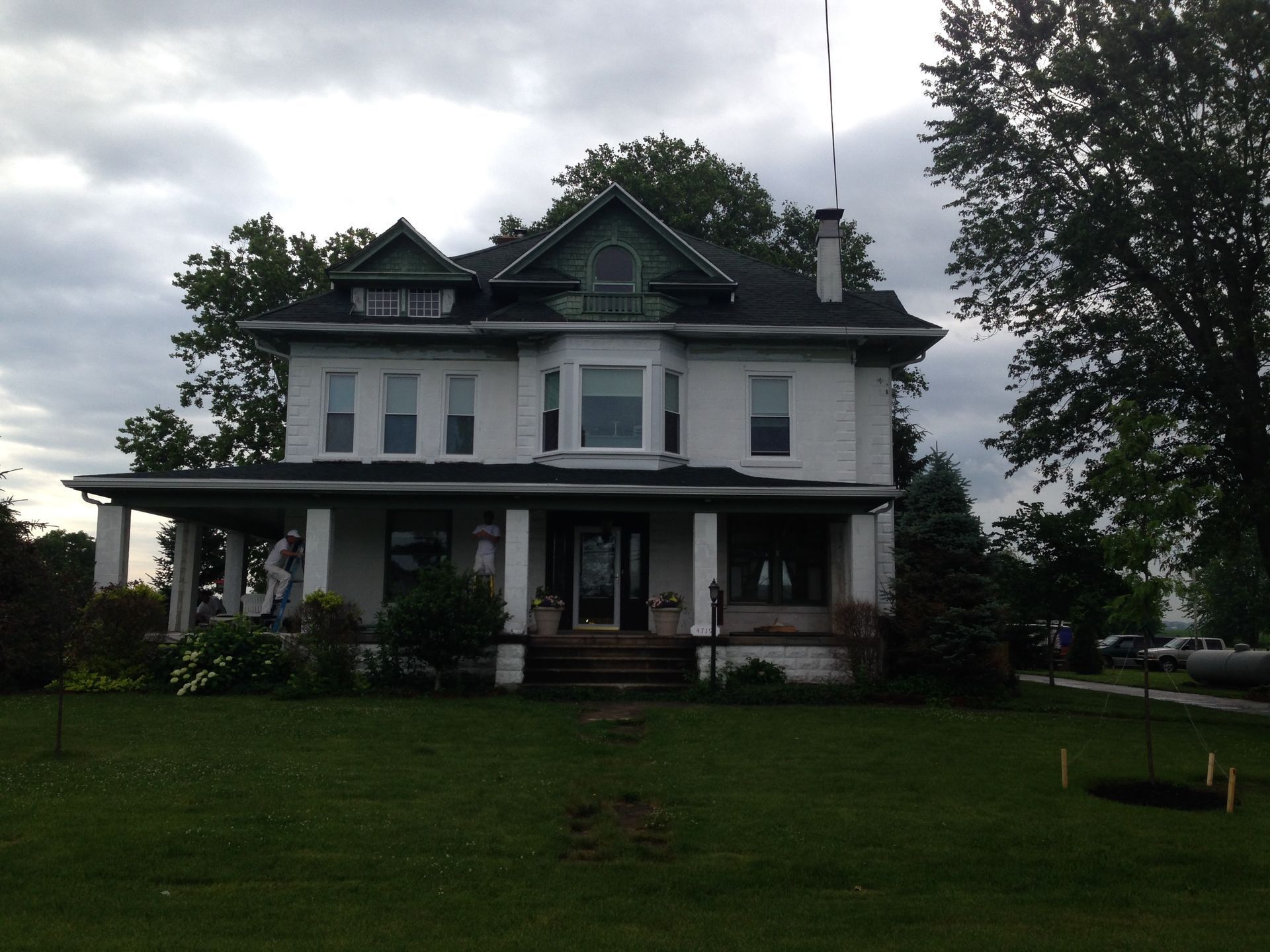 Large white two-story house with a porch, set on a green lawn under a cloudy sky.