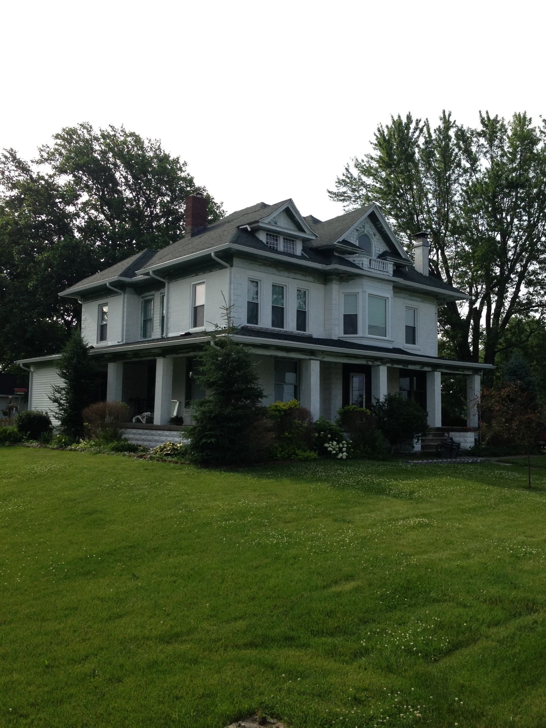 Large white farmhouse with wraparound porch on a grassy hill, framed by tall trees.