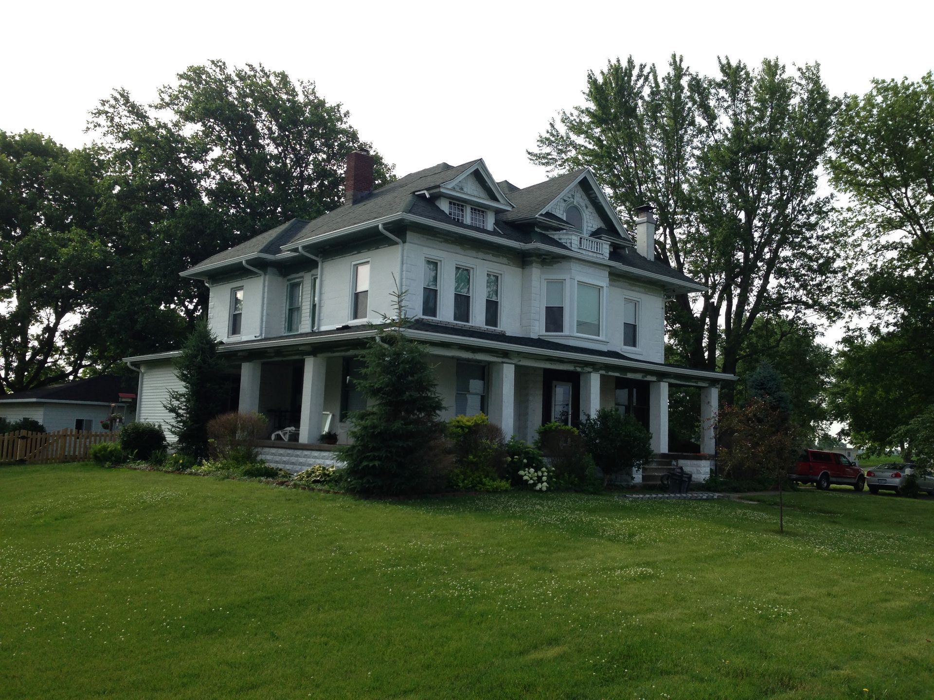 Large white farmhouse with a covered porch on a grassy hill, surrounded by tall trees.