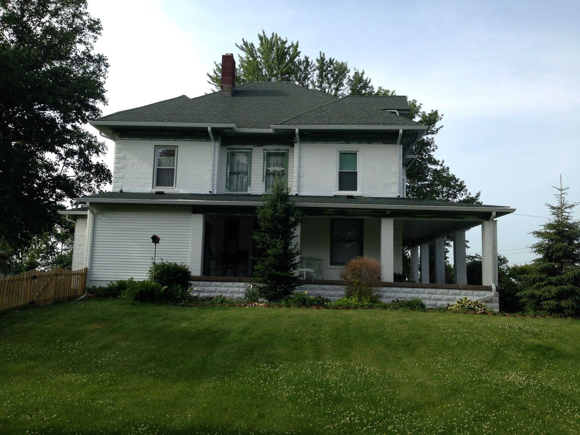 White two-story house with a green roof, covered porch, and large grassy front yard under cloudy sky