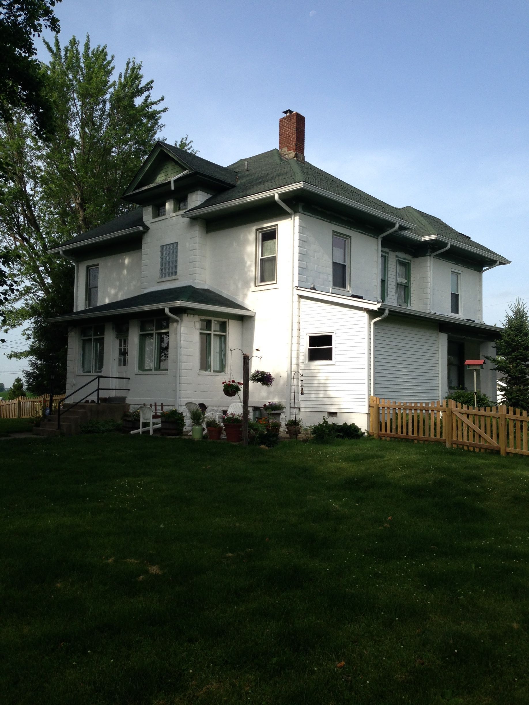 White two-story house with a brick chimney, dark roof, and grassy front yard under trees