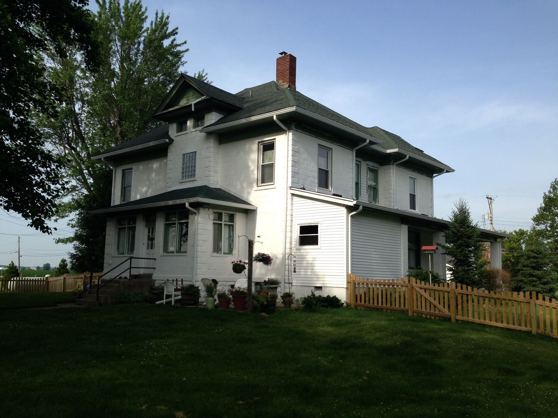 White two-story house with green trim and a red chimney on a grassy lawn behind a wooden fence