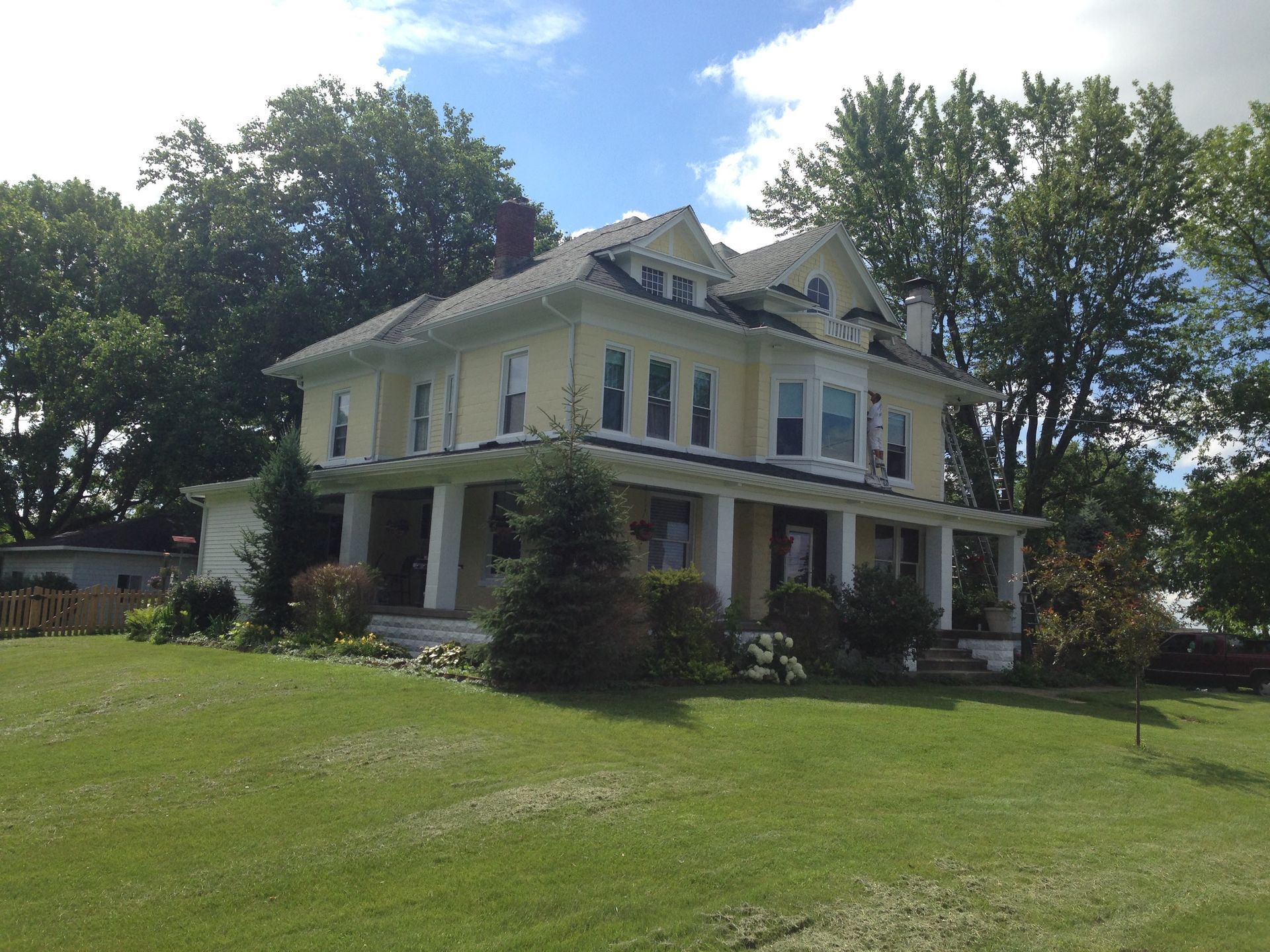 Two-story yellow Victorian house with white trim on a grassy lawn under a partly cloudy sky
