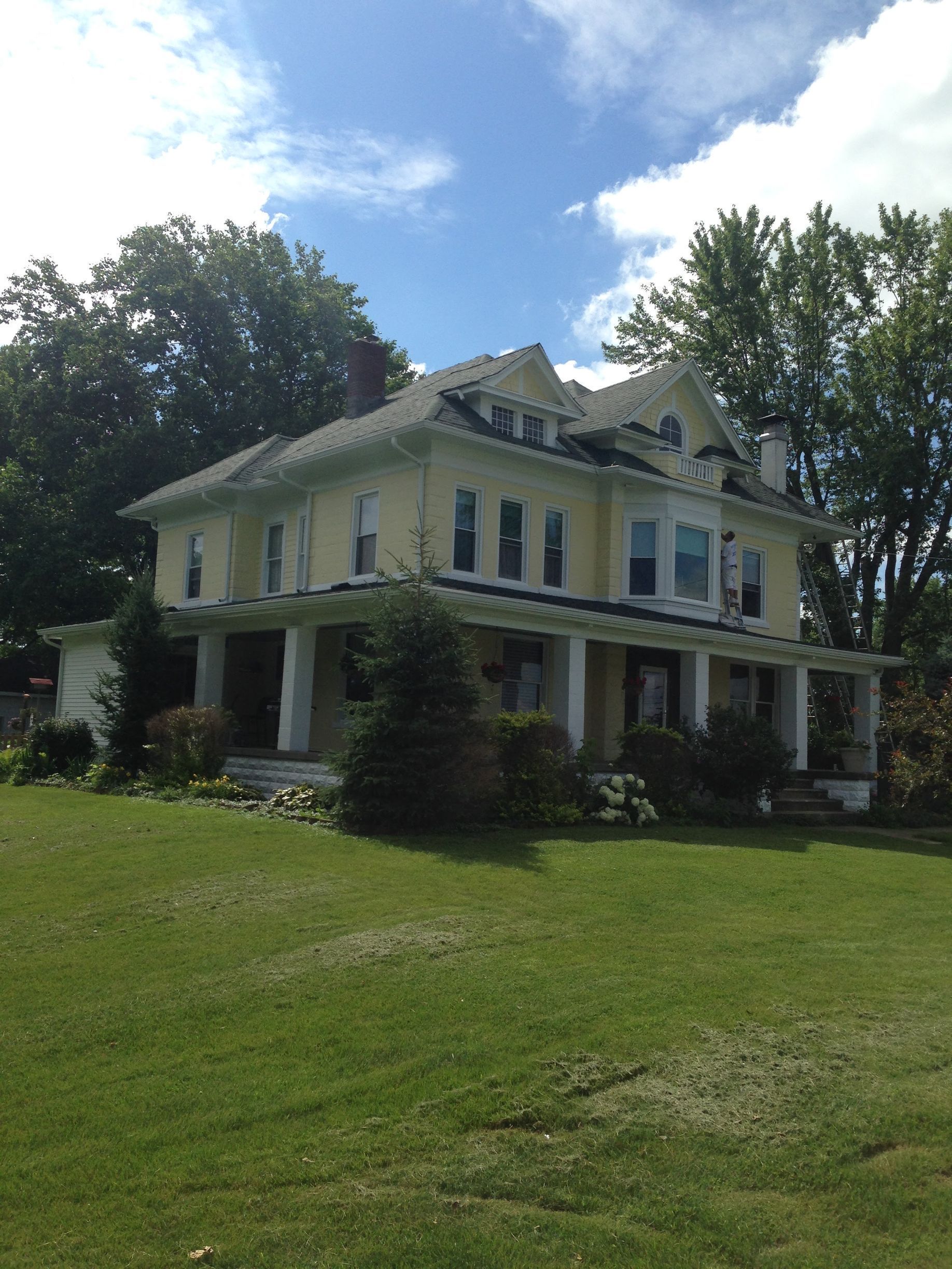 Yellow two-story house with white trim and porch, surrounded by green lawn and trees under a blue sky.