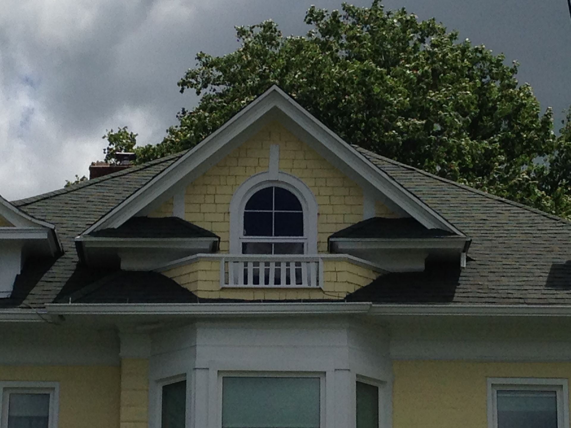 House roof with a dormer window beneath cloudy skies and a leafy tree in the background