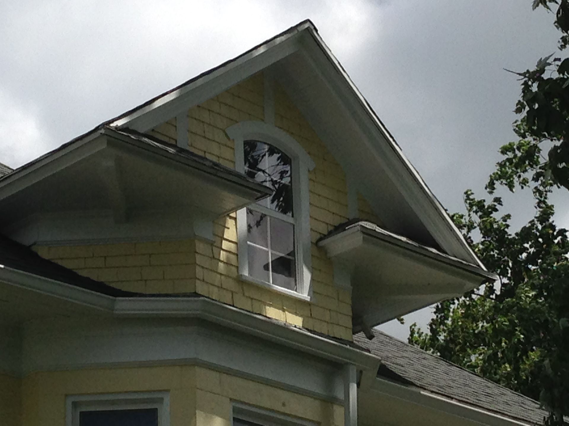 Close-up of a yellow house with white trim, gabled roof, and an arched window against a cloudy sky