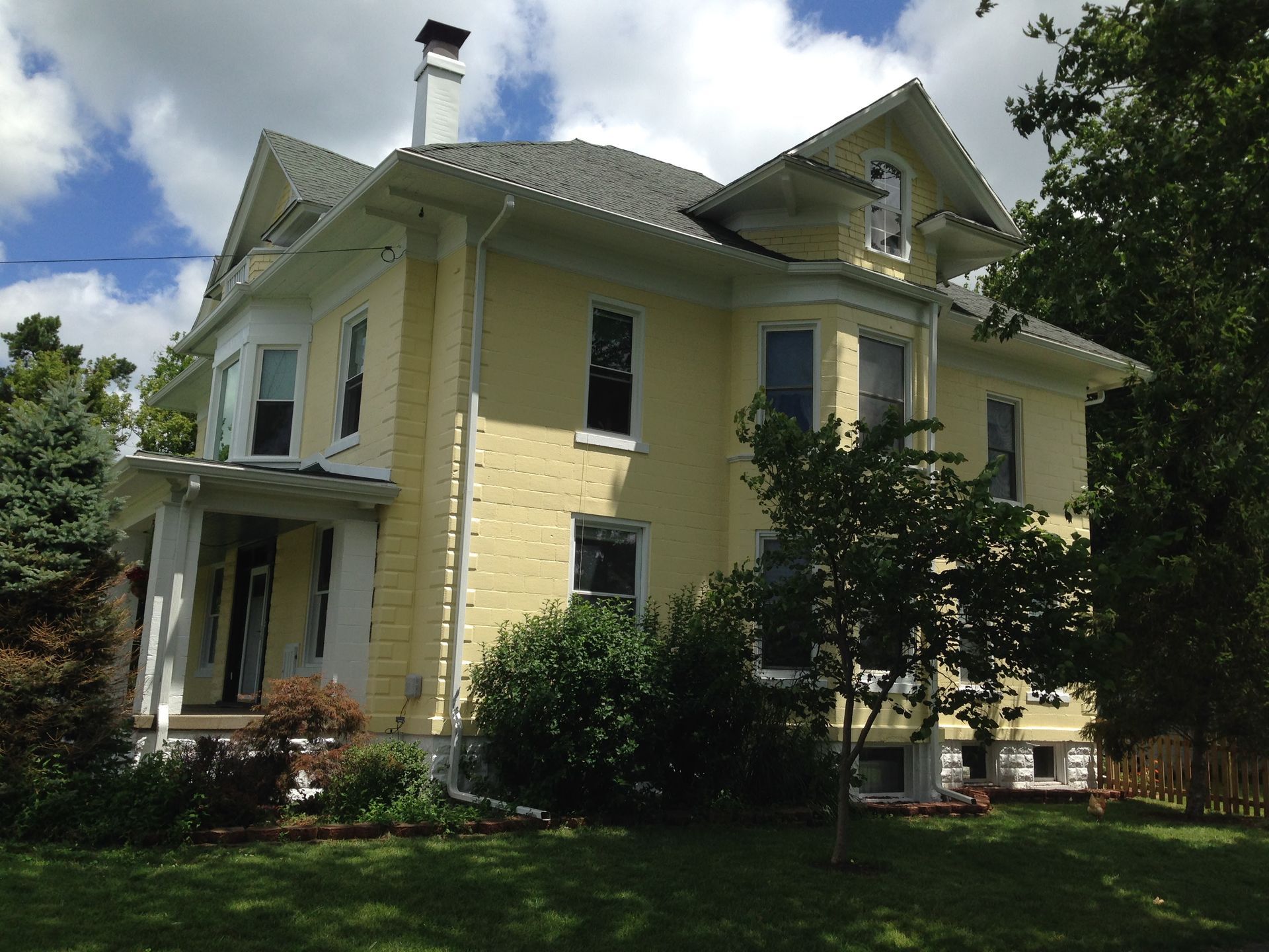 Yellow two-story house with green roof and porch, surrounded by trees and lawn under a cloudy sky