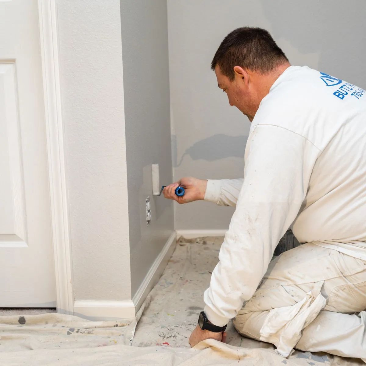 Man kneeling and painting a gray wall baseboard with a small brush in a room under renovation