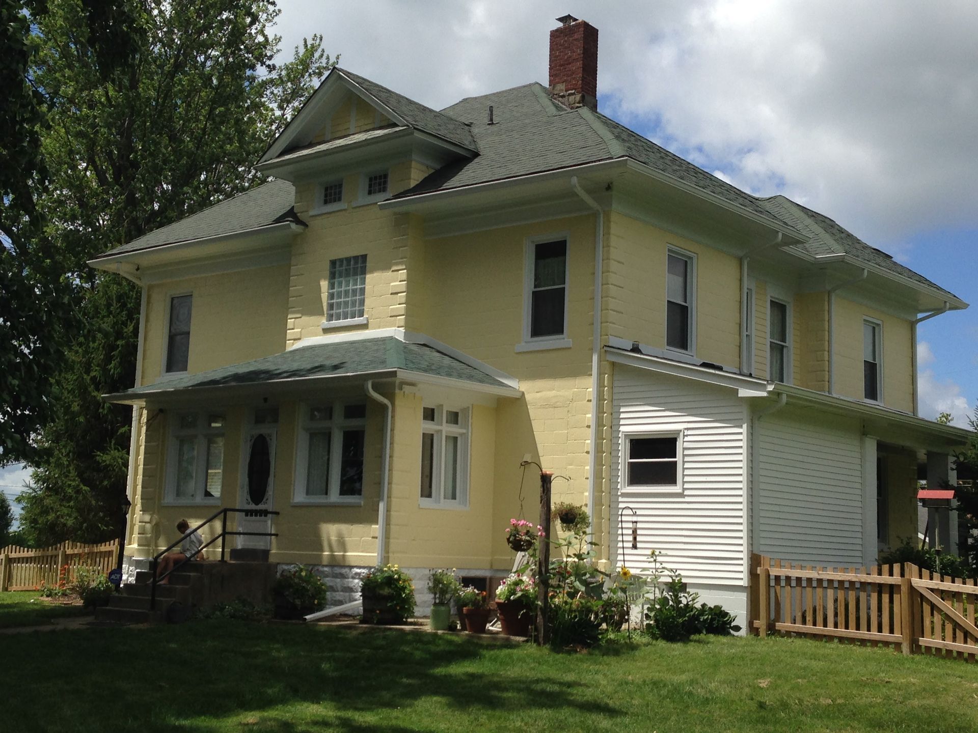 Yellow two-story house with a green roof, front porch, and small fenced yard on a sunny day