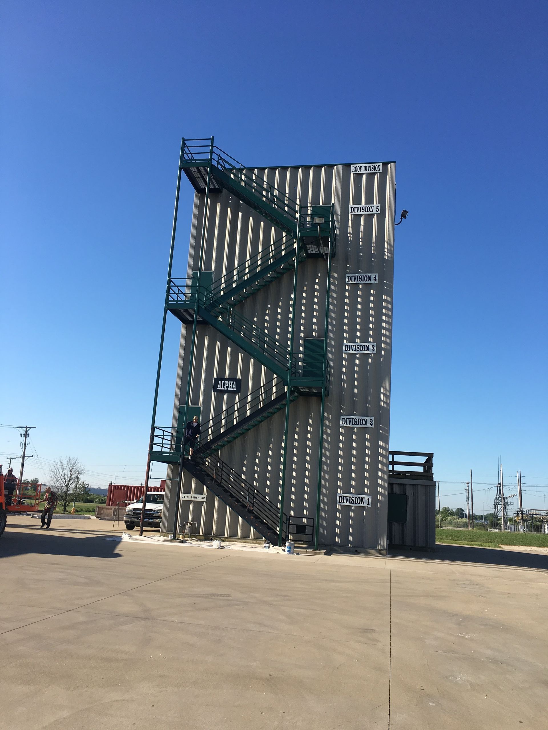 Tall metal climbing wall with green platforms on a concrete lot under a clear blue sky.
