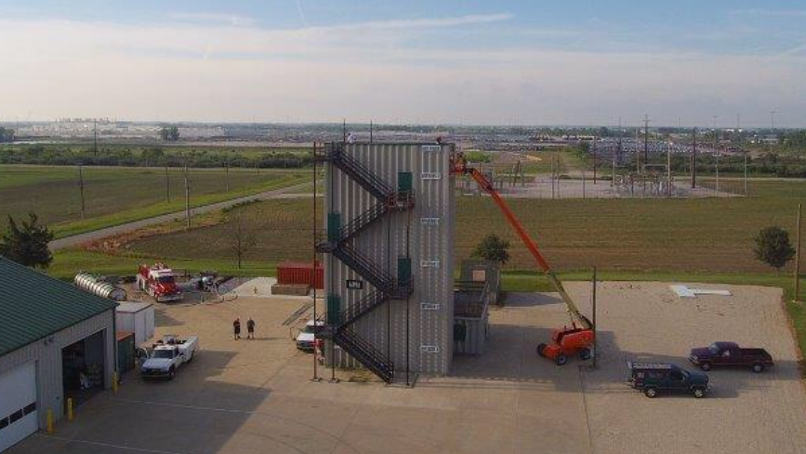 Outdoor industrial training structure with ladders and a boom lift in an open field, viewed from above