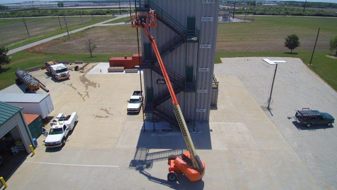 Orange boom lift reaching up beside a large industrial silo in a concrete yard with parked vehicles.