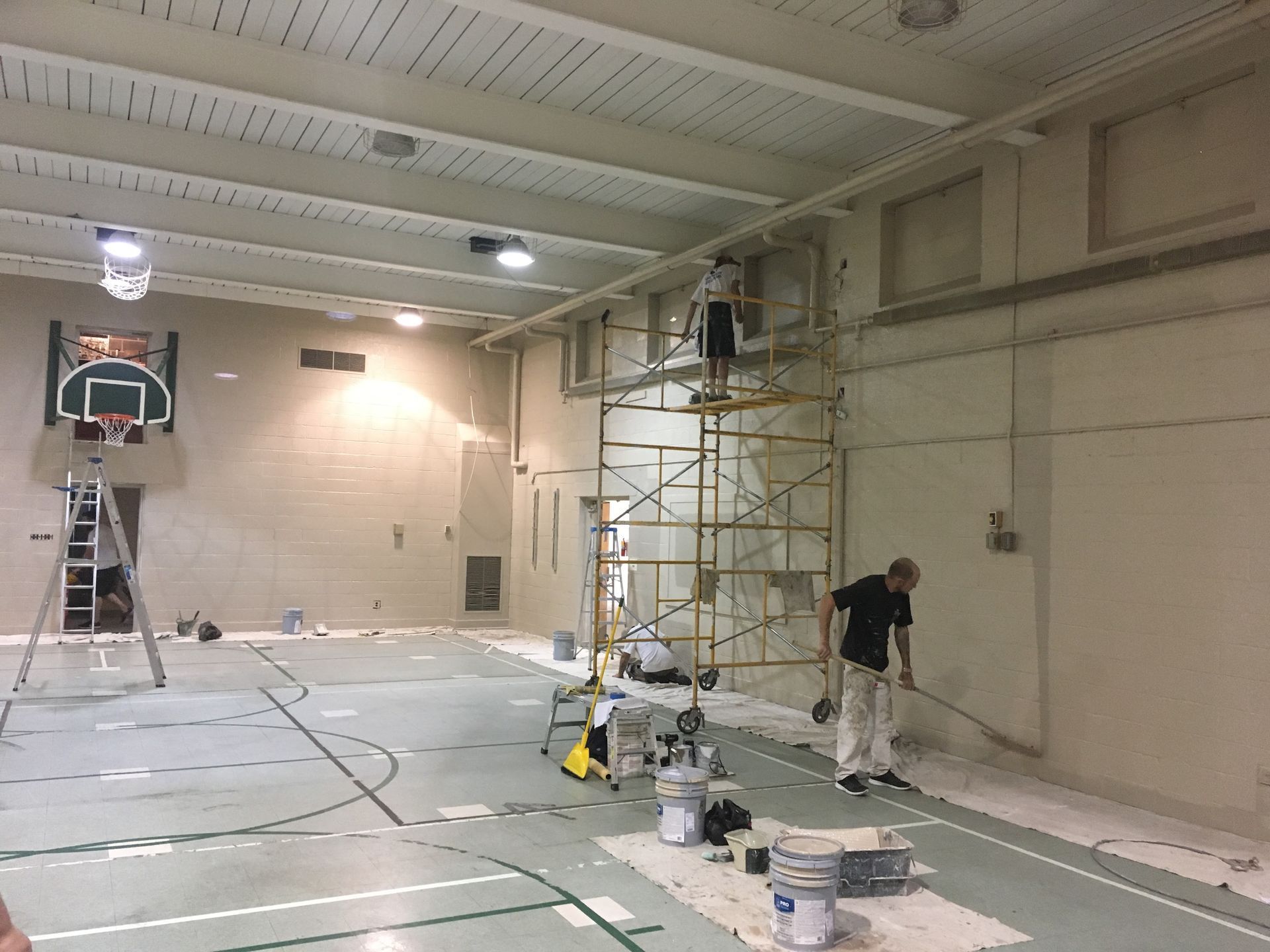 Indoor gym under renovation with scaffolding, ladders, paint buckets, and a worker near the wall.