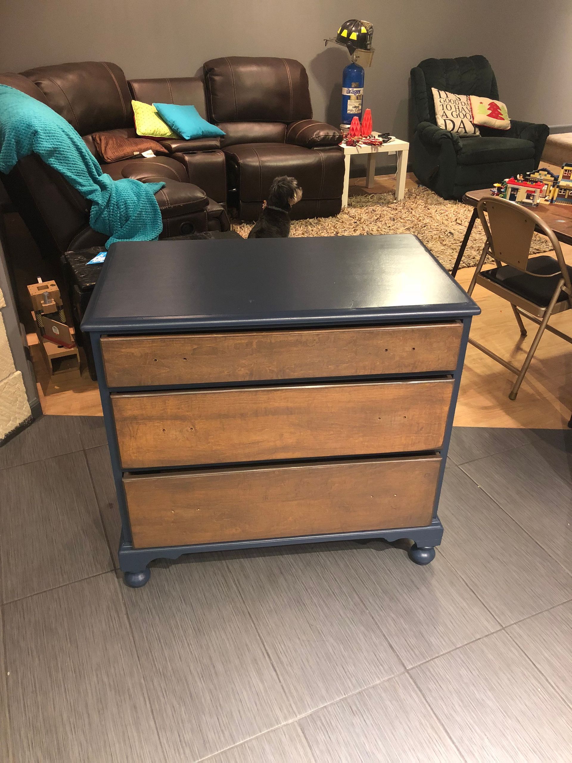 Wooden three-drawer dresser on casters in a living room with leather chairs and a patterned rug.