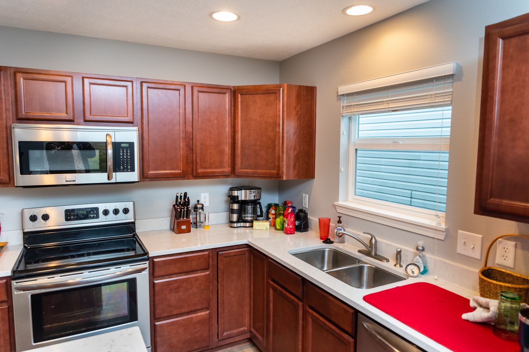 Compact kitchen with wood cabinets, stainless stove and microwave, white countertops, and a sink by a window with blinds.