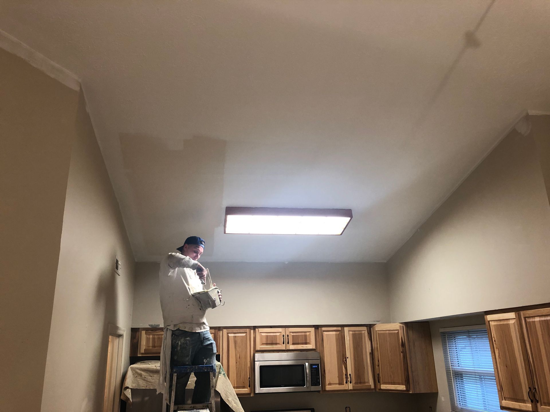 Person standing on a kitchen counter working near the ceiling light above wooden cabinets.