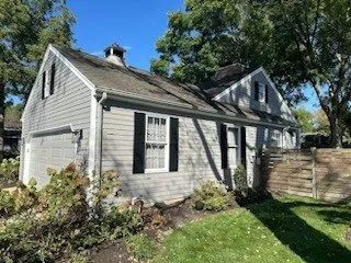 Small gray house with white trim, trees, and a wooden fence in a sunny backyard.