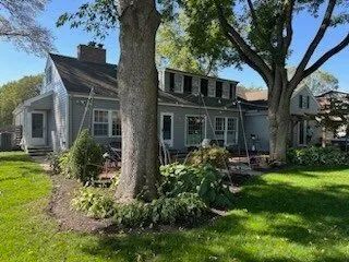 A gray house with white trim and a large lawn shaded by tall trees.