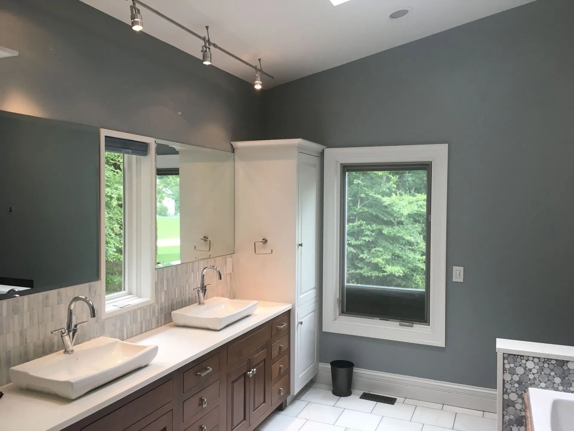 Modern bathroom with double sinks, dark gray walls, and white cabinetry near two windows