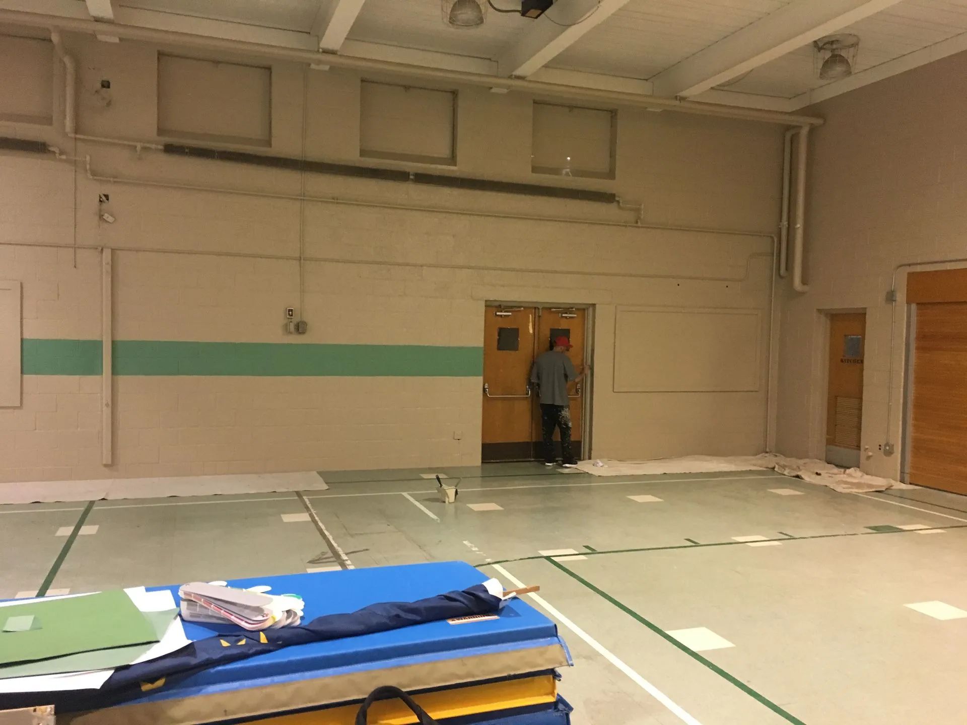 Empty gymnasium with taped floor lines, a person in a doorway, and stacked mats in the foreground