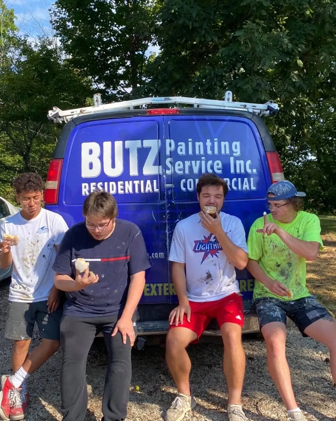Four men sitting on a truck bumper in front of a Butz Painting Service van, smiling and posing outdoors