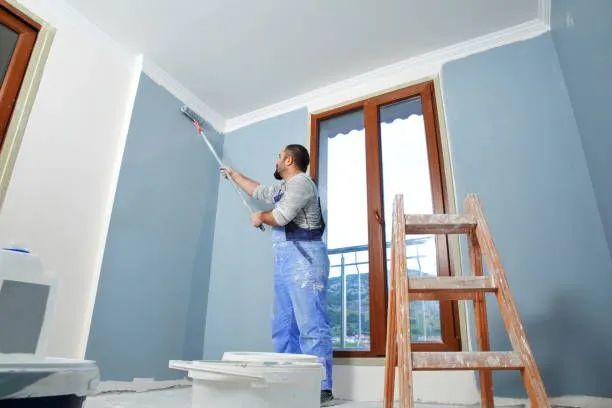 Person painting a light blue room with a roller, ladder and paint supplies visible.