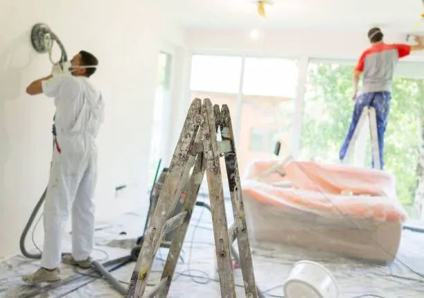 Workers painting a bright room, with a ladder, plastic-covered furniture, and sunlight through windows